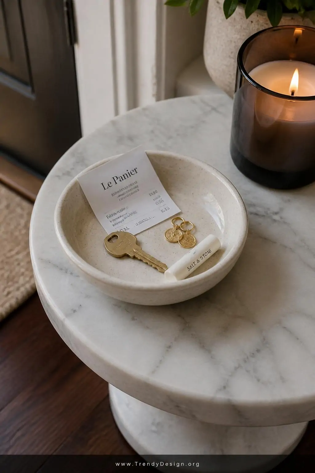 Speckled cream ceramic catch-all bowl resting on a small round marble side table near a front door, holding a brass house key, two gold coin earrings, a chapstick, and a single folded receipt, warm candlelight glowing beside it in a smoked glass holder, rich dark walnut flooring beneath, extreme closeup shot, photorealistic interior design.