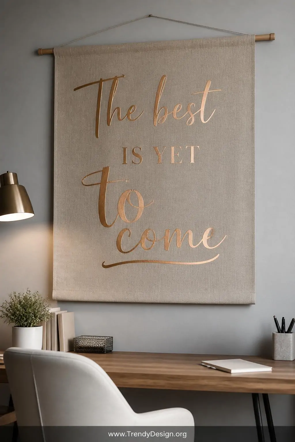 Stretched linen fabric wall hanging on a wooden dowel with large rose gold metallic vinyl quote lettering mounted above a minimalist desk, soft warm ambient light, medium shot showing the dramatic metallic sheen against the natural undyed linen backdrop.