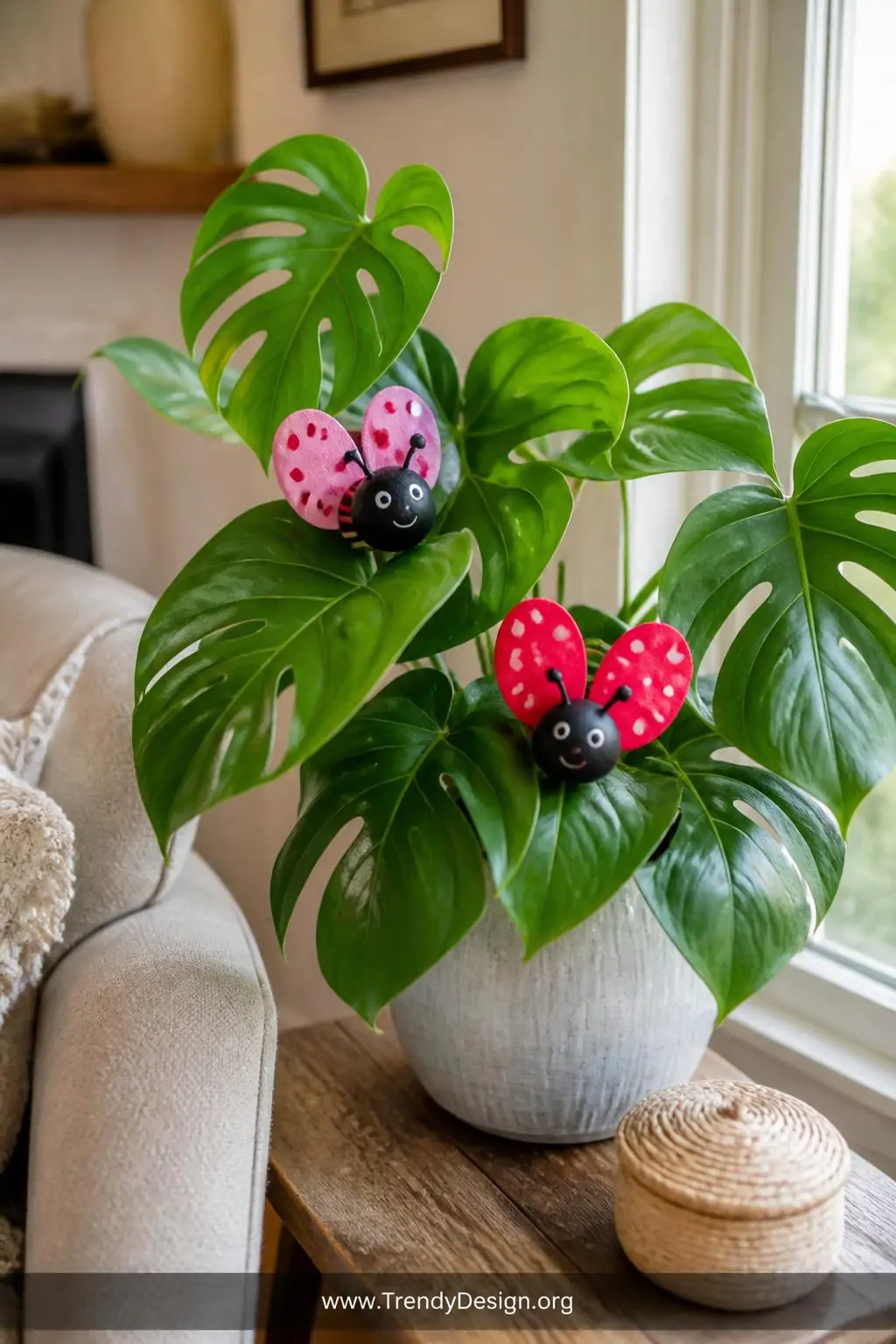 Bright painted salt dough love bug figures with heart-shaped wings in red and pink nestled among the leaves of a potted monstera plant on a sunlit living room side table, cheerful natural morning light, medium shot.