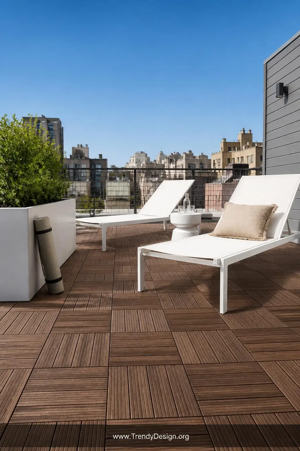 Wood-grain embossed rubber deck tiles in warm walnut-effect finish covering a bright rooftop terrace, interlocking joints visible at edges, lightweight contemporary outdoor loungers in white, rolled yoga mat leaning against a low planter, clear blue sky backdrop, city rooftop context, medium shot, photorealistic interior design.